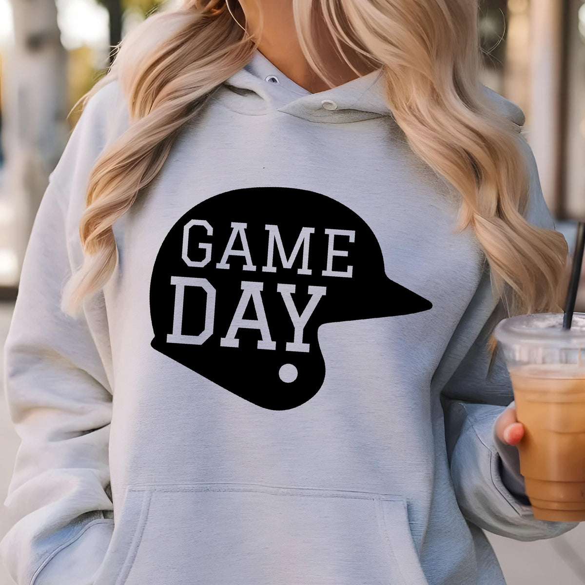 May include: A light blue hooded sweatshirt featuring a black graphic of a baseball helmet with the white text GAME DAY printed inside. The garment includes a front kangaroo pocket, long sleeves, and a drawstring hood with a visible white button.