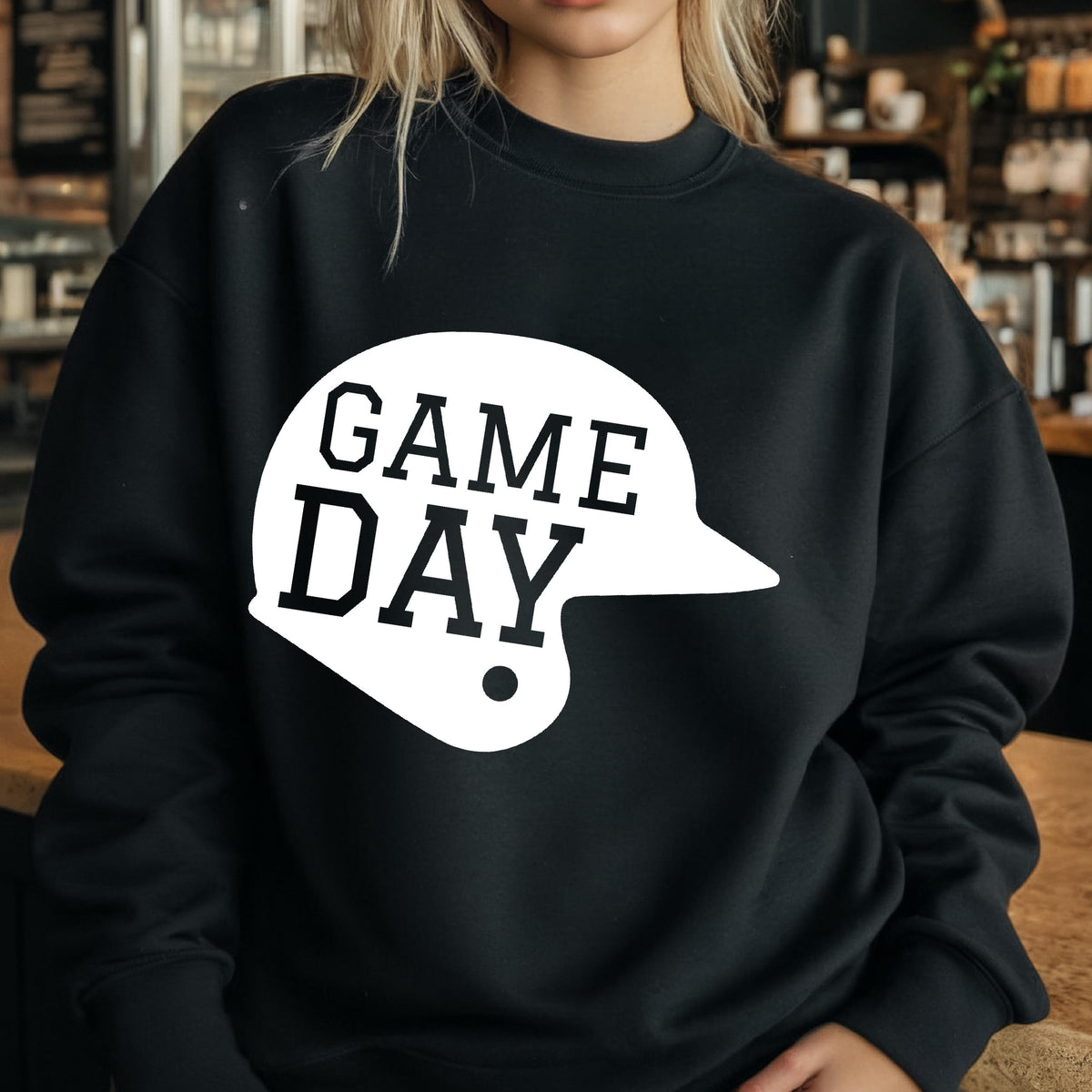 May include: A black long-sleeve crewneck sweatshirt featuring a large white graphic of a baseball helmet silhouette. The text "GAME DAY" is printed in bold black block letters inside the white helmet shape on the front of the garment.