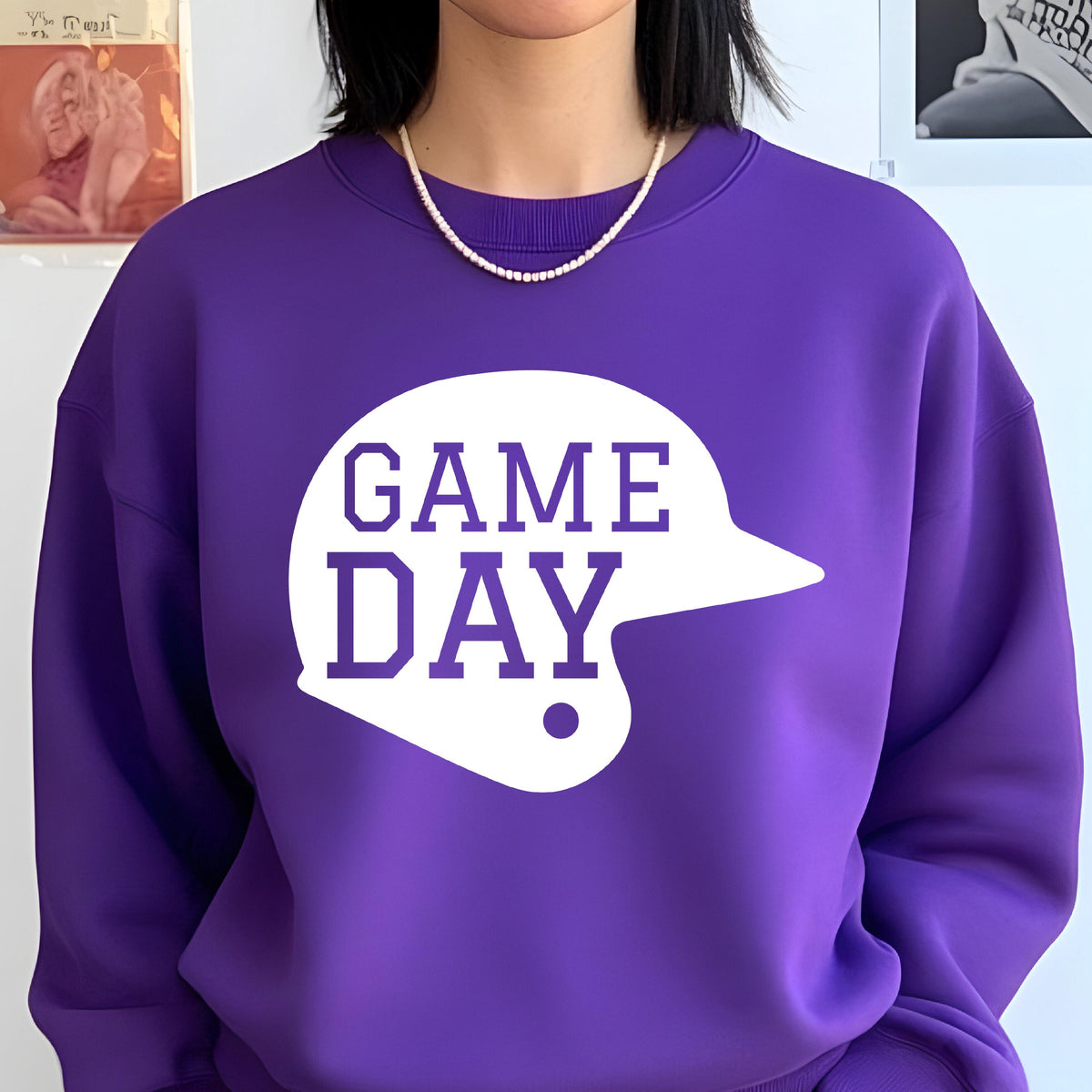 May include: A vibrant purple crewneck sweatshirt featuring a large white graphic of a baseball helmet silhouette. The text GAME DAY is printed in bold purple block letters inside the helmet shape, centered on the chest of the garment.