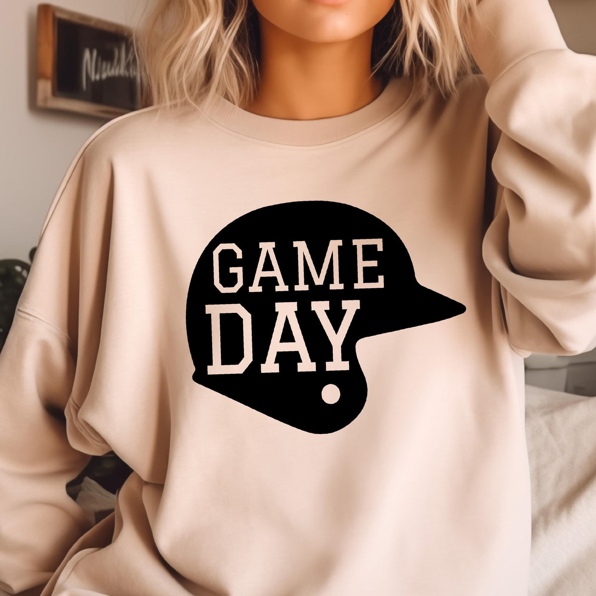 May include: A beige crewneck sweatshirt featuring a black graphic of a baseball helmet with the text GAME DAY printed in a light beige font across the center of the helmet shape. The long-sleeved garment has a simple, solid-colored design.