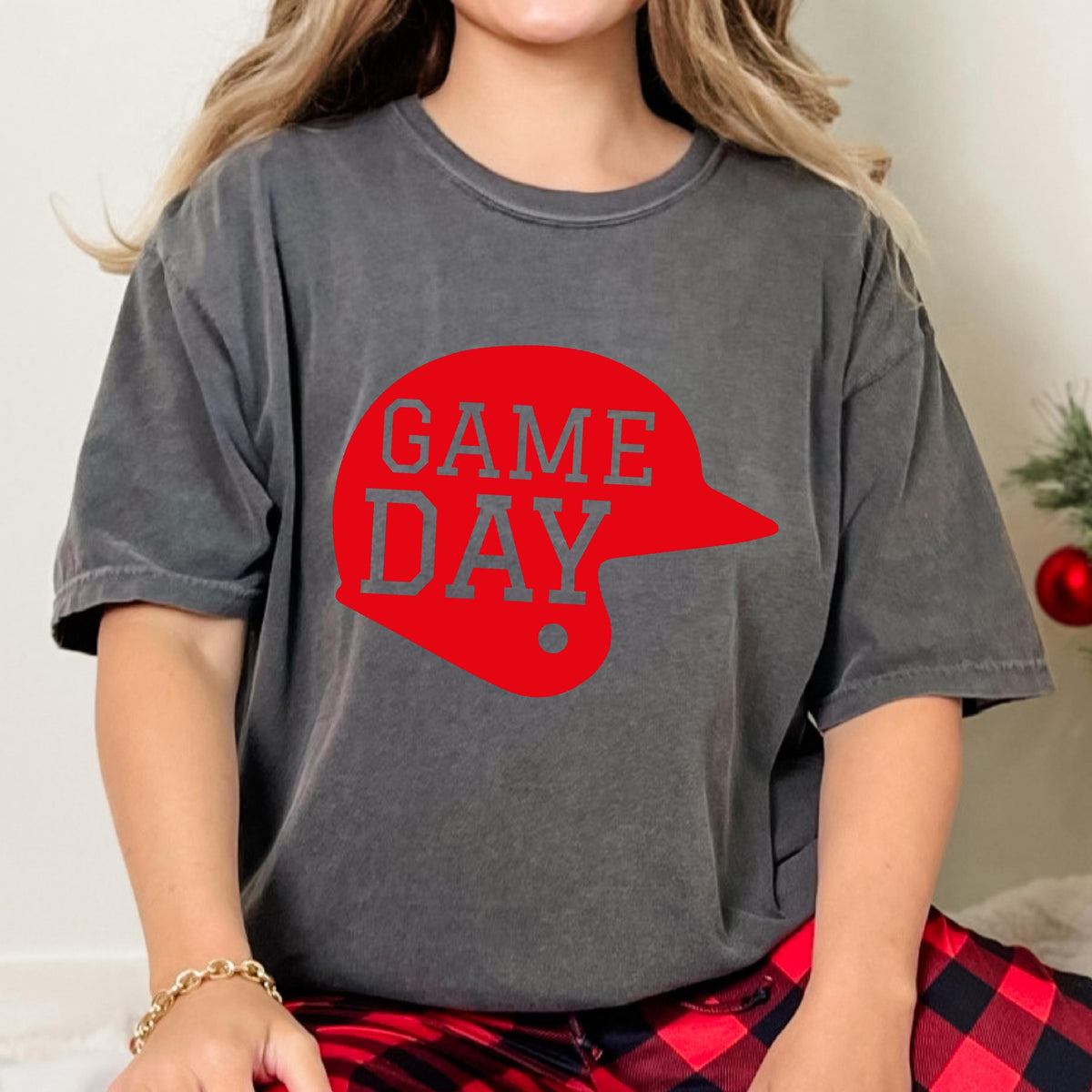 May include: A charcoal grey short-sleeve t-shirt featuring a red graphic of a baseball helmet silhouette with the text GAME DAY printed in grey block letters across the center of the helmet design on the front of the garment.