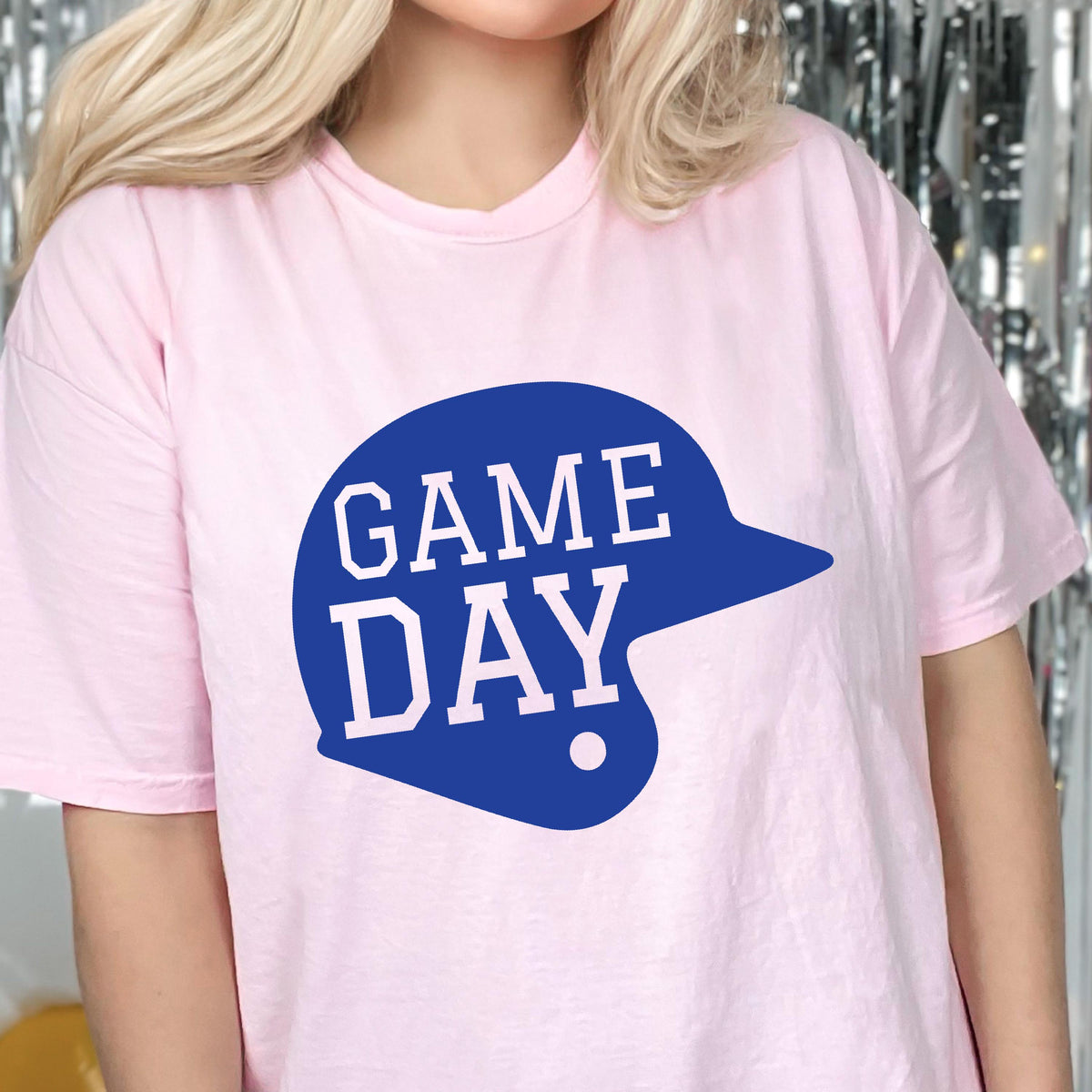 May include: A light pink short-sleeved t-shirt featuring a large, solid blue graphic of a baseball helmet. The text "GAME DAY" is printed in white, block-style capital letters across the center of the blue helmet graphic on the front.