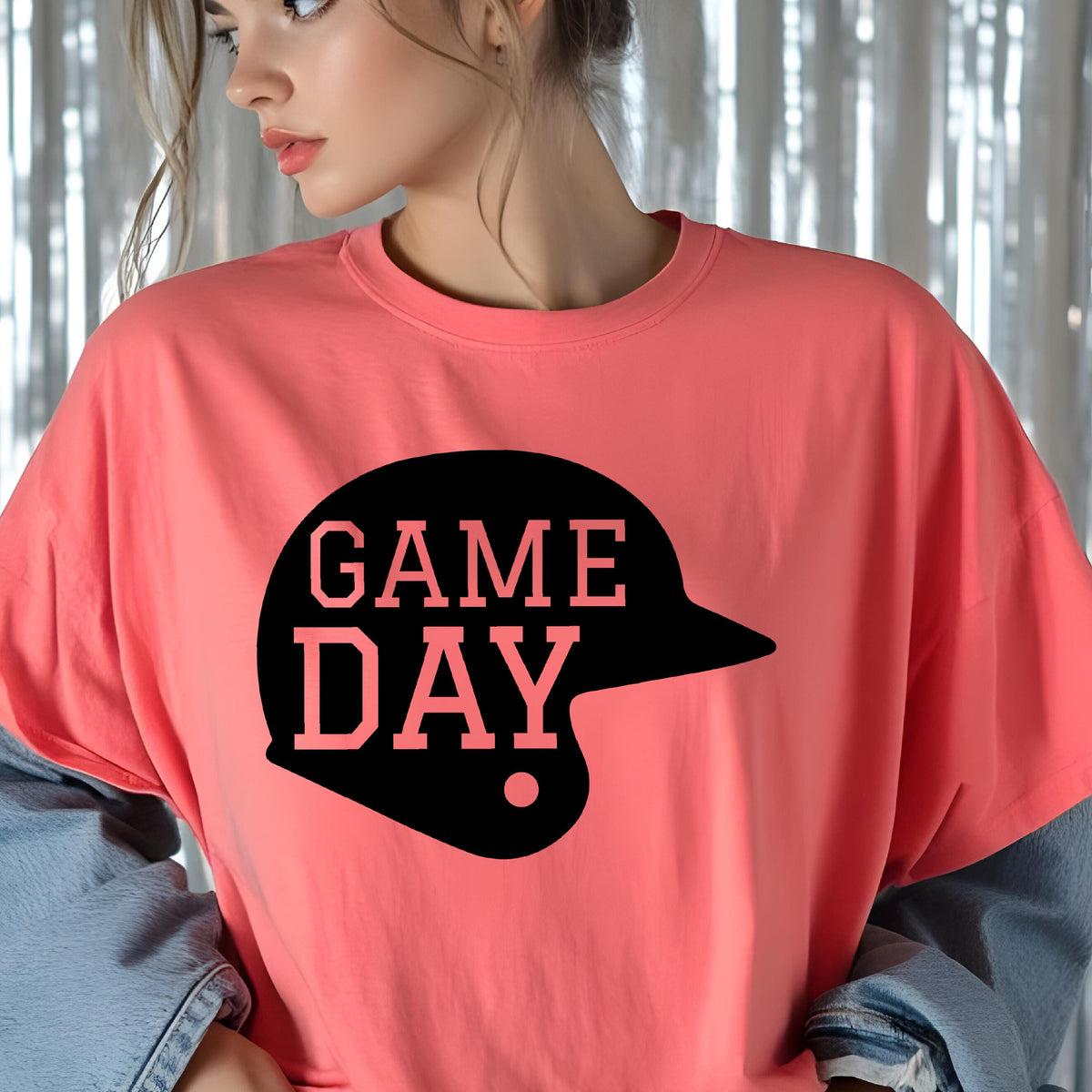 May include: A coral-colored short-sleeve t-shirt featuring a black graphic of a baseball helmet silhouette. The text GAME DAY is printed in a bold, block-style font across the center of the helmet graphic in a light coral color.