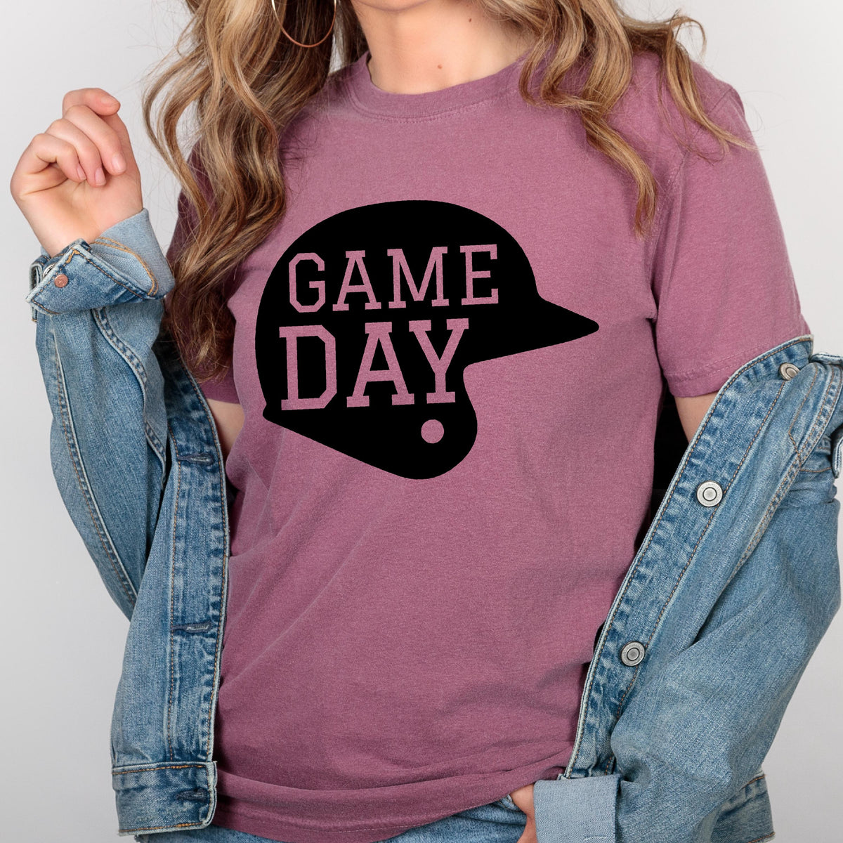 May include: A mauve short-sleeved t-shirt featuring a black graphic of a baseball helmet with the text GAME DAY printed in a light pink font inside the helmet shape, worn under a blue denim jacket with visible metal buttons and stitching.