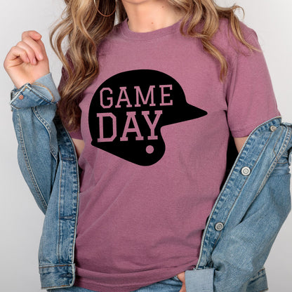 May include: A mauve short-sleeved t-shirt featuring a black graphic of a baseball helmet with the text GAME DAY printed in a light pink font inside the helmet shape, worn under a blue denim jacket with visible metal buttons and stitching.