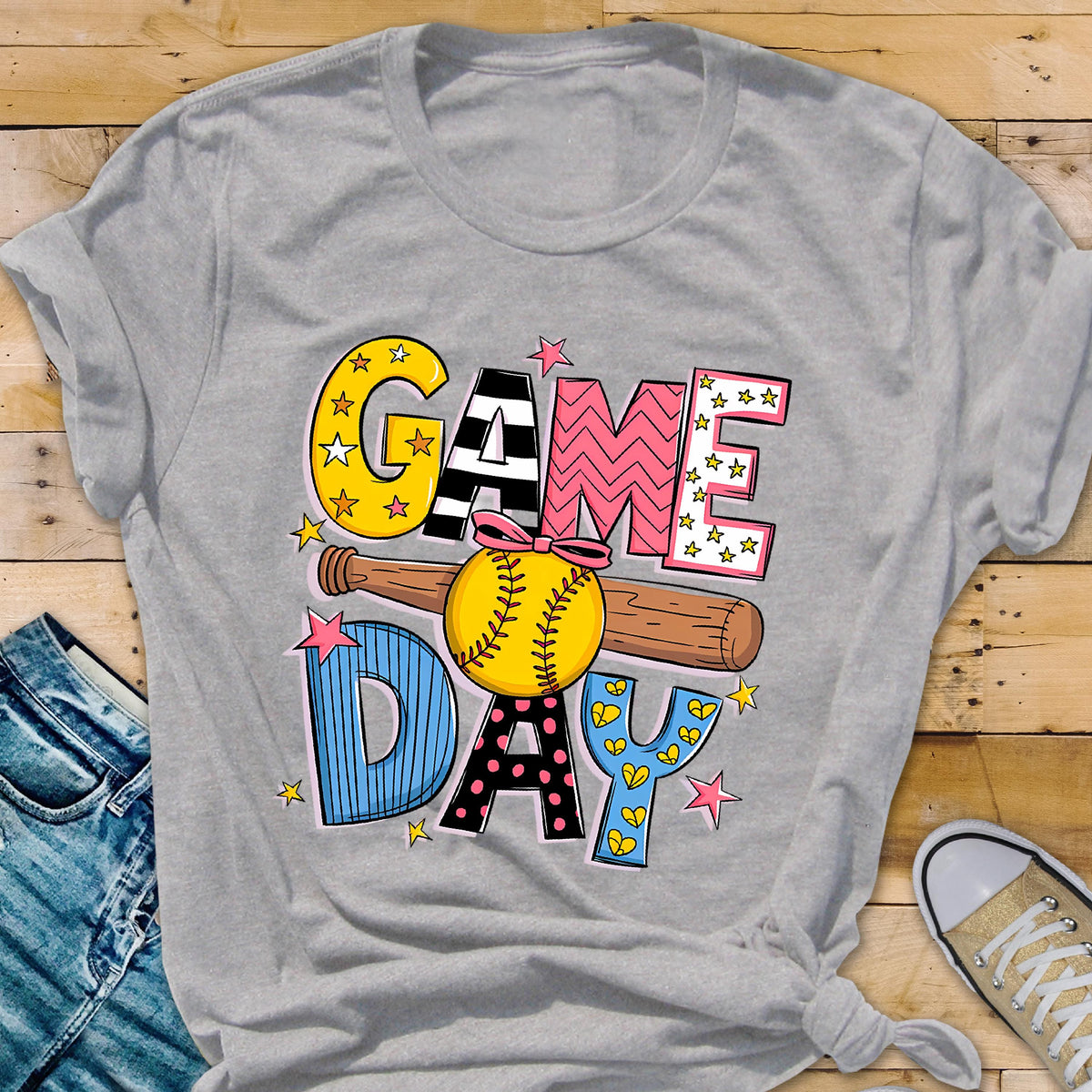 May include: A gray t-shirt with a colorful graphic design that says "GAME DAY" with a baseball and bat. The graphic is made up of different colors and patterns, including stars, polka dots, and stripes.