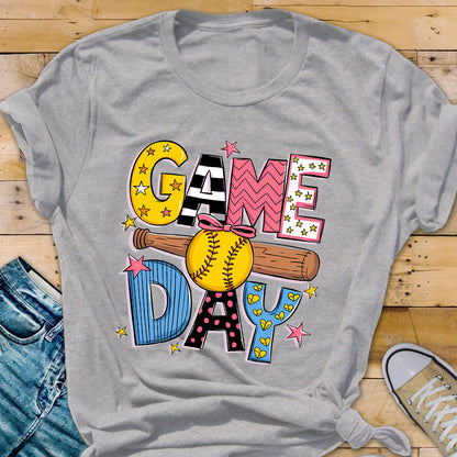 May include: A gray t-shirt with a colorful graphic design that says "GAME DAY" with a baseball and bat. The graphic is made up of different colors and patterns, including stars, polka dots, and stripes.