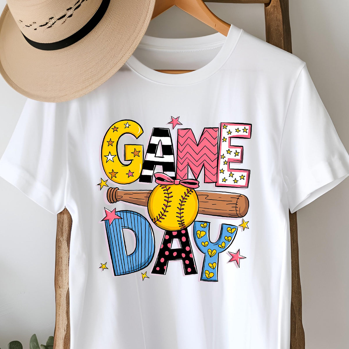 May include: White t-shirt with a colorful graphic design that says "GAME DAY" with a baseball and bat. The letters are decorated with stars, stripes, polka dots, and hearts.