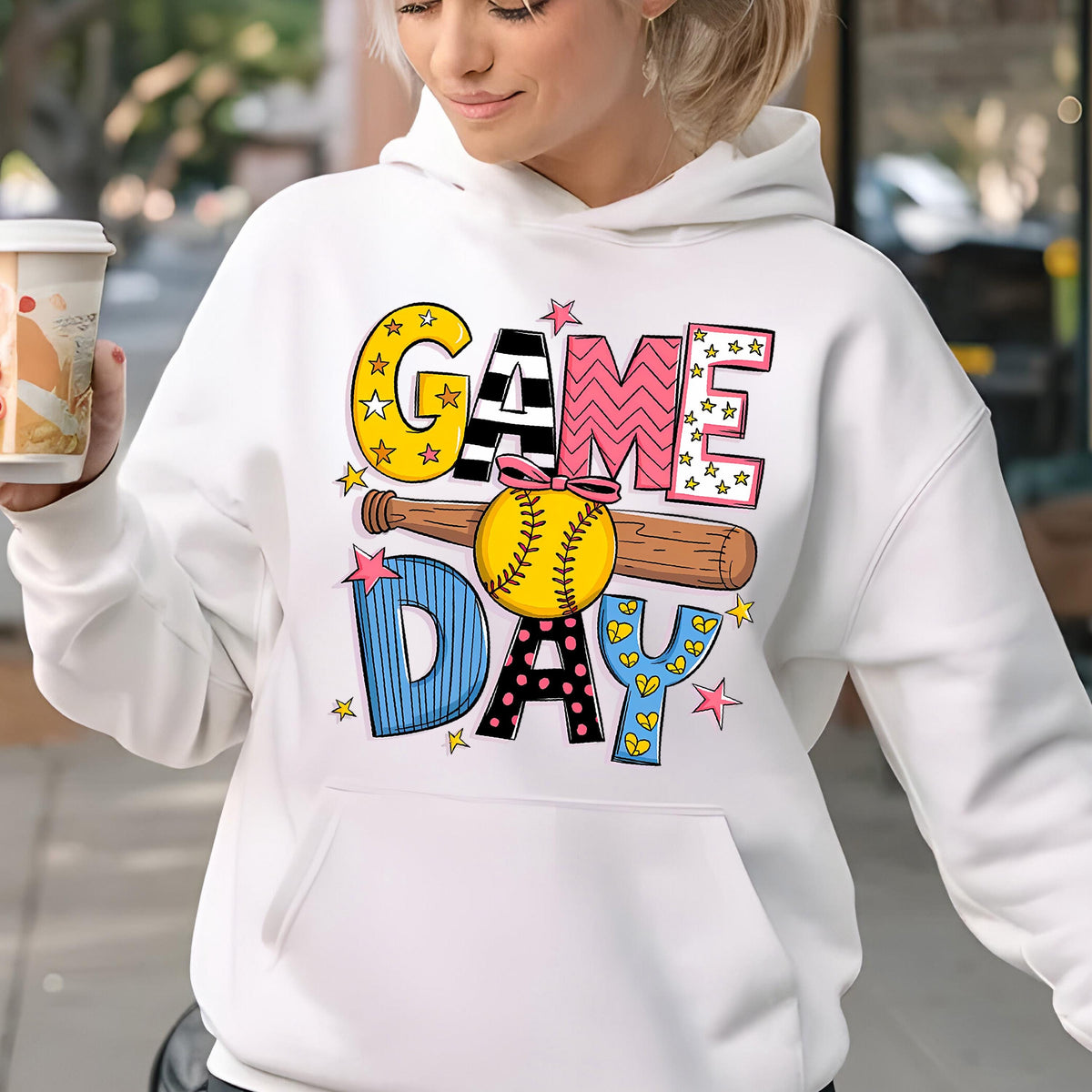 May include: A white hooded sweatshirt featuring a graphic print that reads GAME DAY in colorful, patterned block letters. A brown wooden baseball bat and a yellow softball with a pink bow are centered between the words, accented by small pink stars.