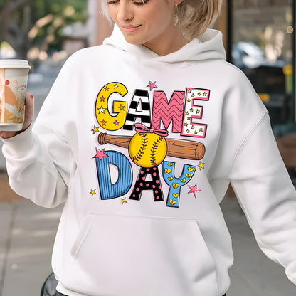 May include: A white hooded sweatshirt featuring a graphic print that reads GAME DAY in colorful, patterned block letters. A brown wooden baseball bat and a yellow softball with a pink bow are centered between the words, accented by small pink stars.