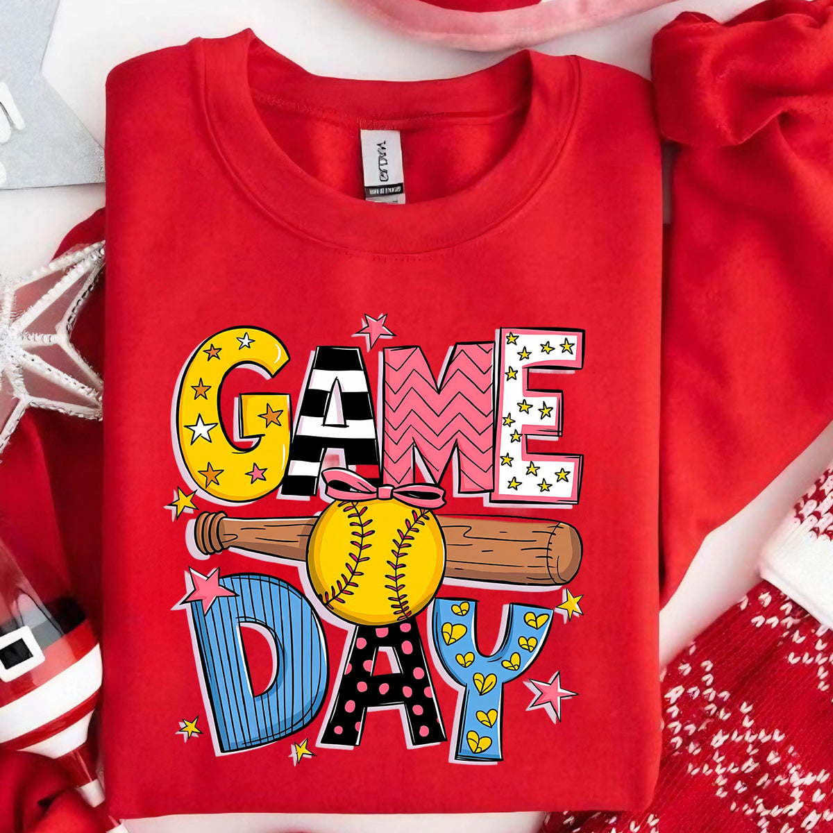 May include: A bright red crewneck sweatshirt featuring a graphic print with the text GAME DAY. The design includes a yellow softball with a pink bow, a wooden baseball bat, and colorful letters decorated with stars, stripes, and polka dots.
