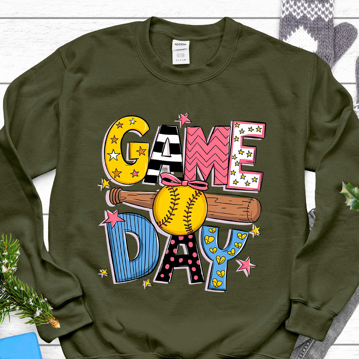 May include: An olive green crewneck sweatshirt featuring a graphic print with the text GAME DAY. The design includes a yellow softball with a pink bow, a brown baseball bat, and colorful letters decorated with stars, stripes, and polka dots.