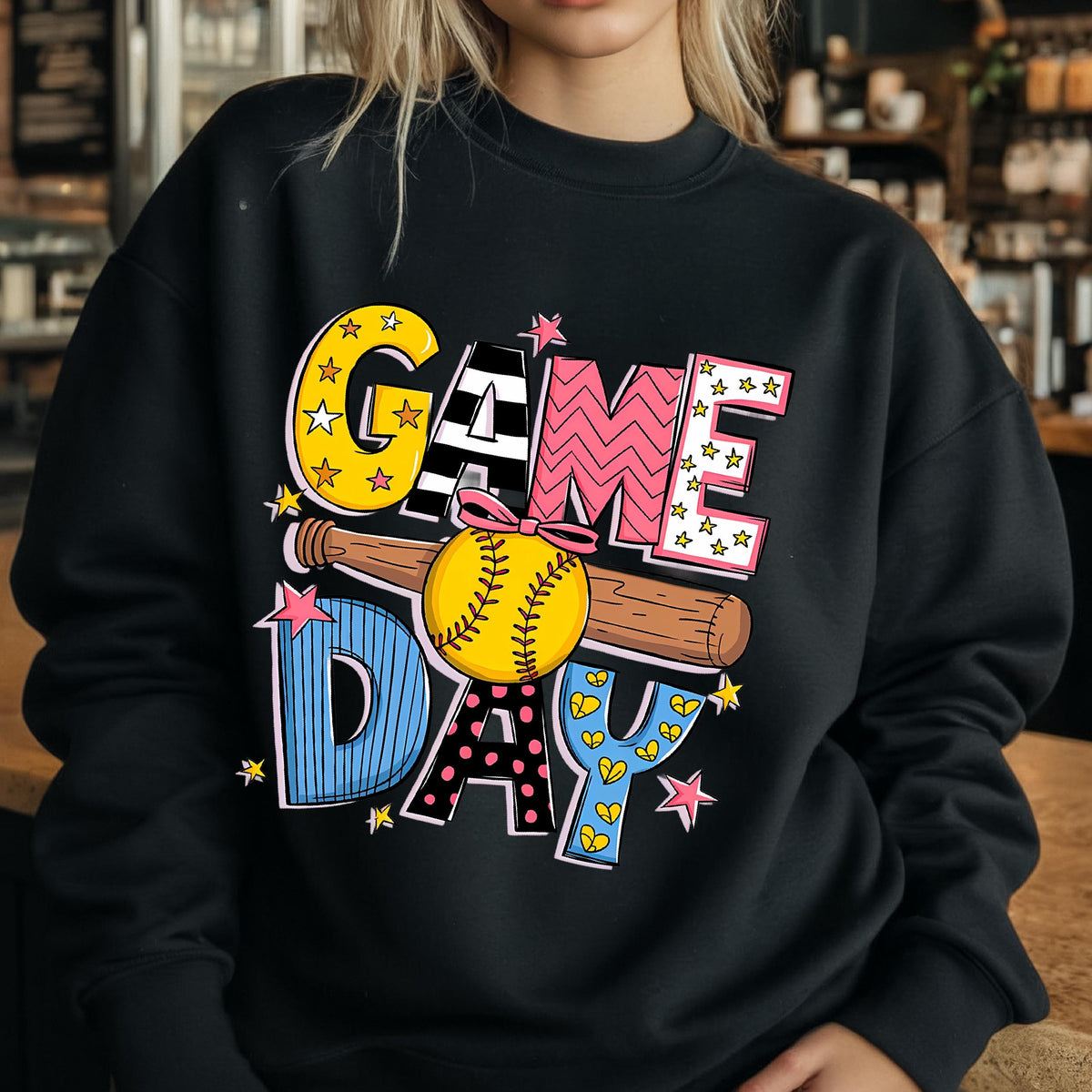 May include: A black crewneck sweatshirt featuring a graphic print that reads GAME DAY in large, multi-patterned letters. A brown wooden baseball bat and a yellow softball with a pink bow are positioned horizontally across the center of the text.