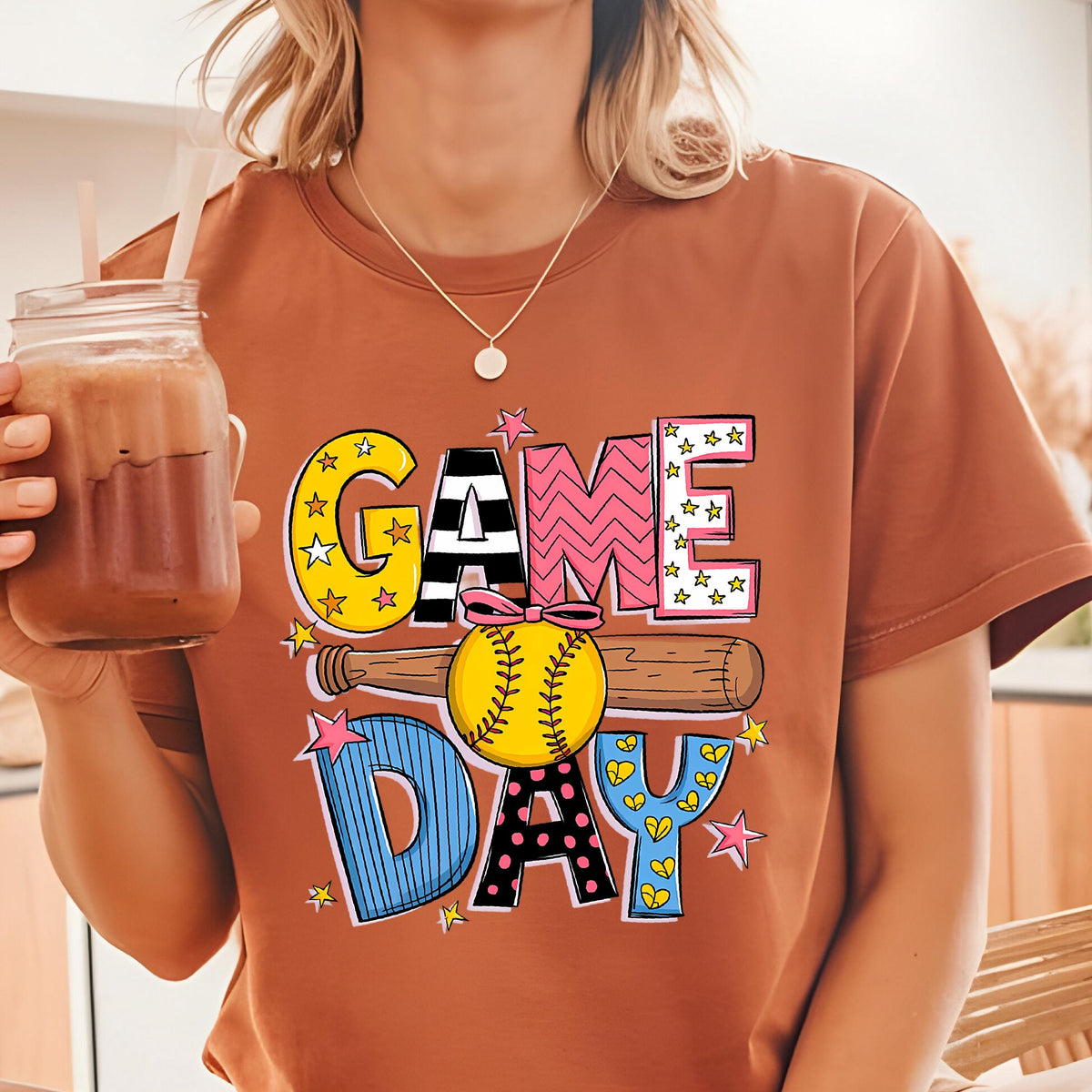 May include: A brown t-shirt with a colorful graphic design that says "GAME DAY" with a baseball and bat. The graphic features stars, hearts, and polka dots.