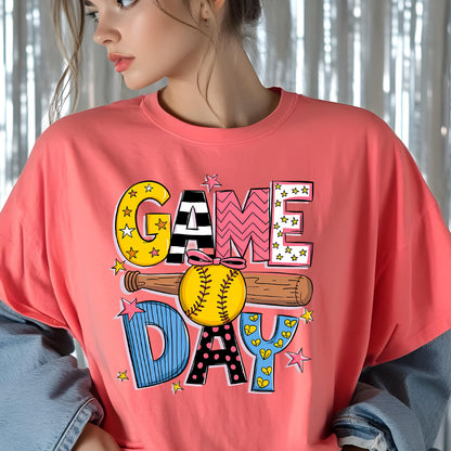 May include: A pink t-shirt with a colorful graphic design that says "GAME DAY" with a baseball and bat. The letters are decorated with stars, hearts, and polka dots.