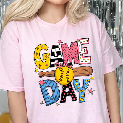 May include: Pink t-shirt with a colorful graphic design that says "GAME DAY" with a baseball and bat. The letters are decorated with stars, stripes, and polka dots.