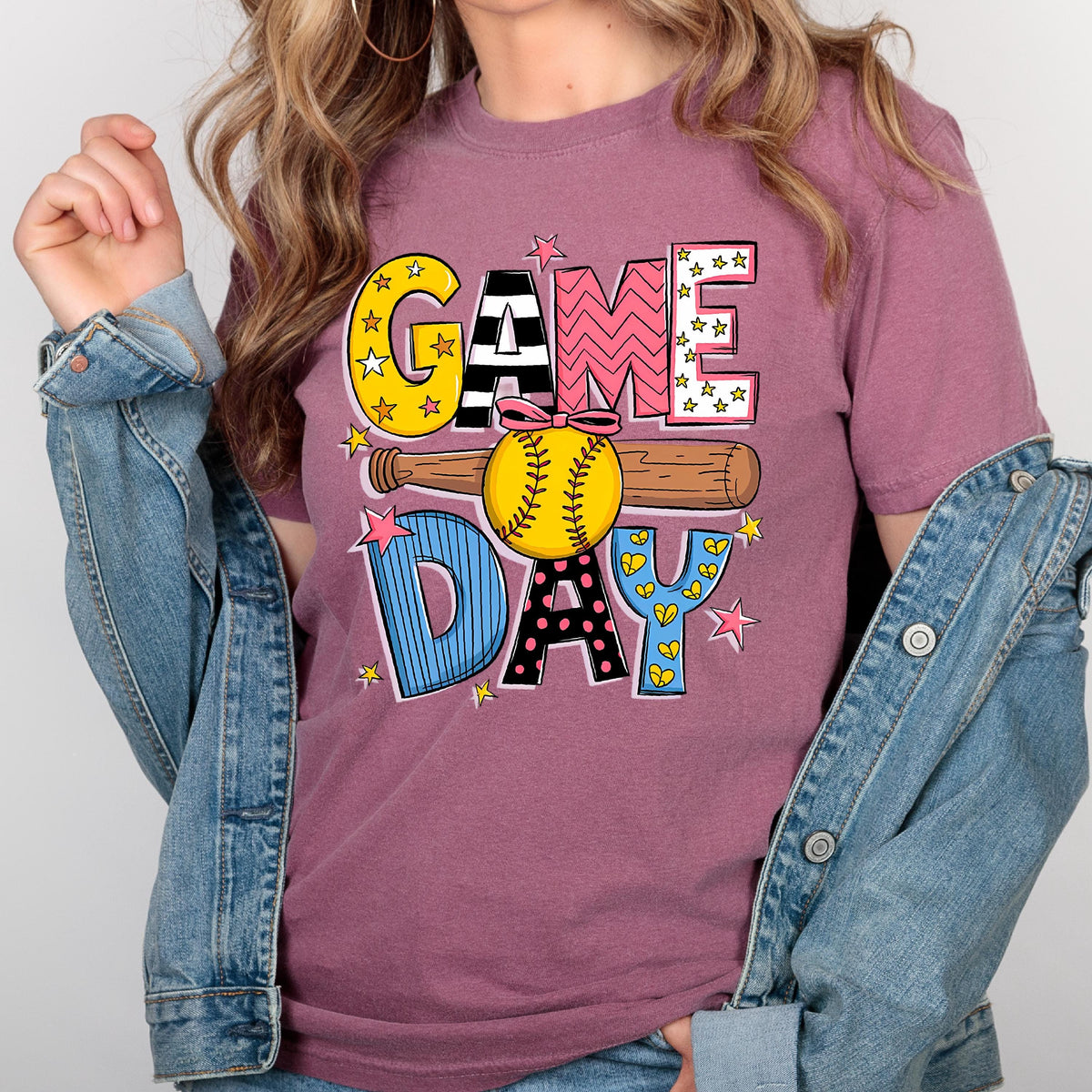 May include: A pink t-shirt with the words "GAME DAY" in colorful, patterned letters. A baseball and bat are behind the letters. The shirt is being worn by a person.