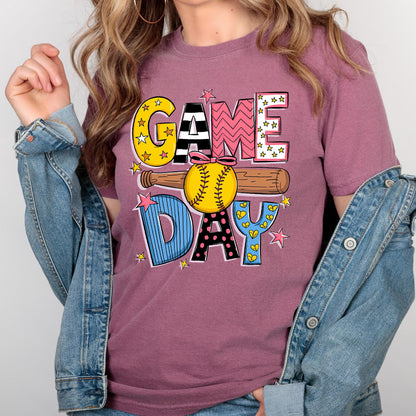 May include: A pink t-shirt with the words "GAME DAY" in colorful, patterned letters. A baseball and bat are behind the letters. The shirt is being worn by a person.