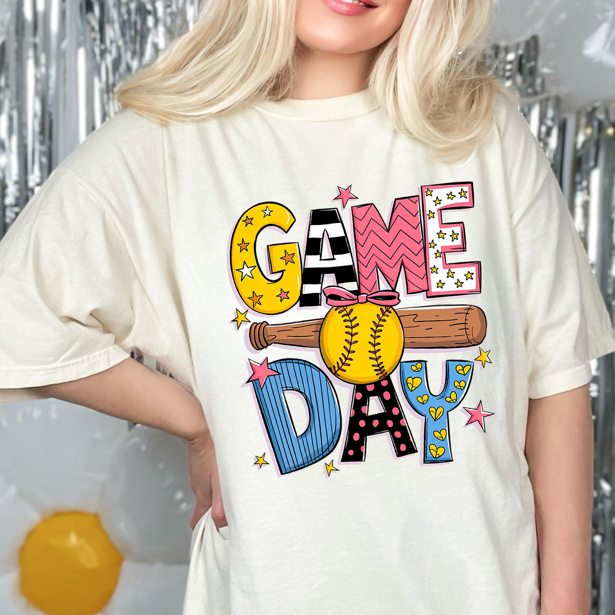 May include: A white t-shirt with a colorful graphic design that says "GAME DAY" with a baseball and bat. The letters are in different colors and patterns.