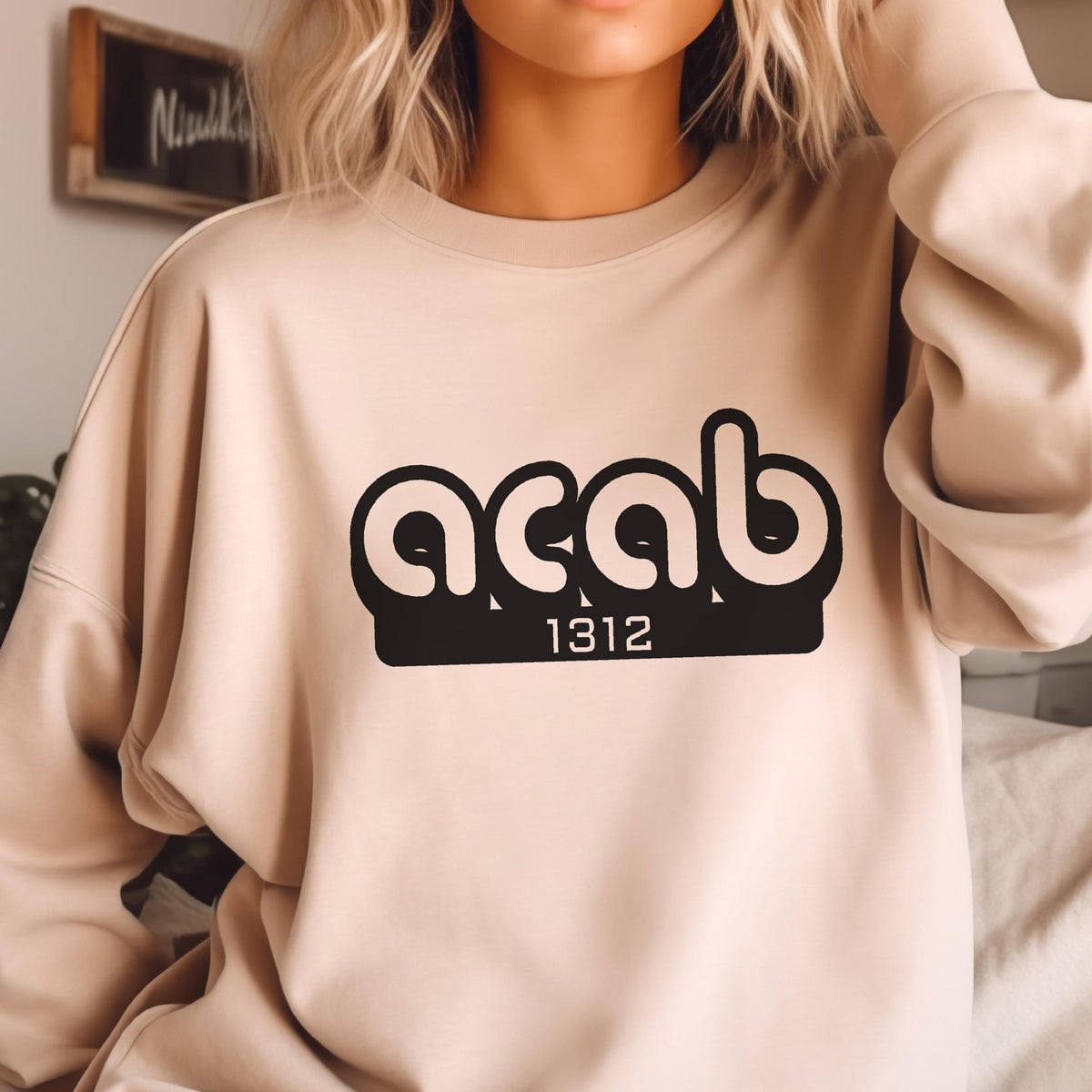 May include: A beige sweatshirt with a black graphic print of the letters "ACAB" in a rounded font with the number "1312" underneath.