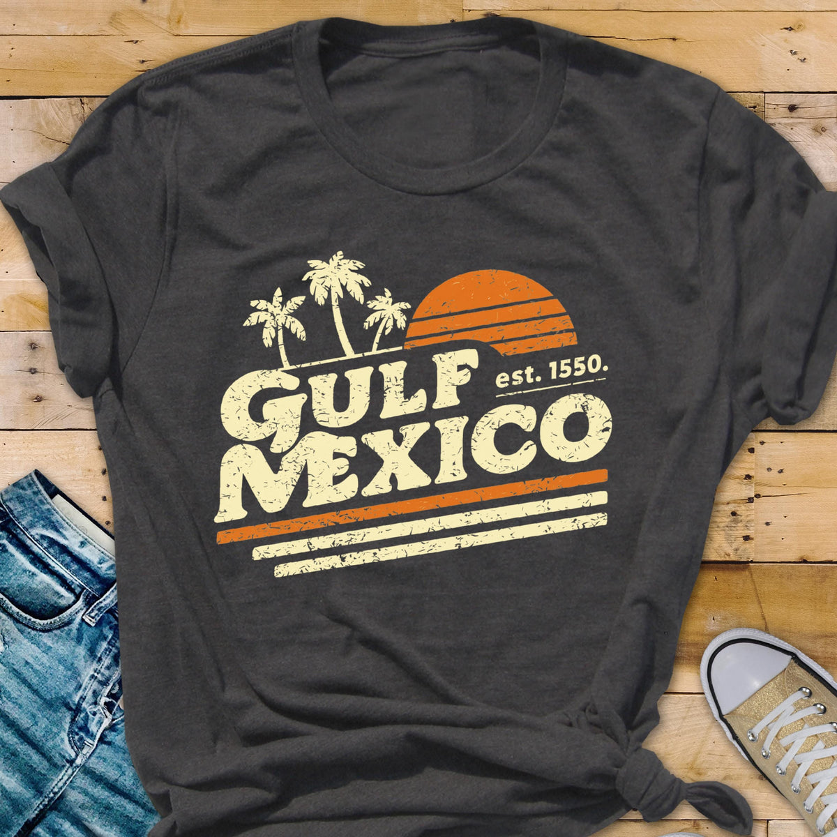 May include: Dark gray t-shirt with a retro design featuring the words "GULF MEXICO" in a vintage font, palm trees, and an orange sun. The text "est. 1550" is also included. The shirt has a classic crew neck and rolled sleeves.