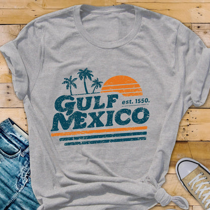 May include: A gray t-shirt featuring the words "GULF MEXICO" in teal, with an orange sun and palm trees graphic. The text "est. 1550" is also included. The shirt is a casual style, suitable for everyday wear.