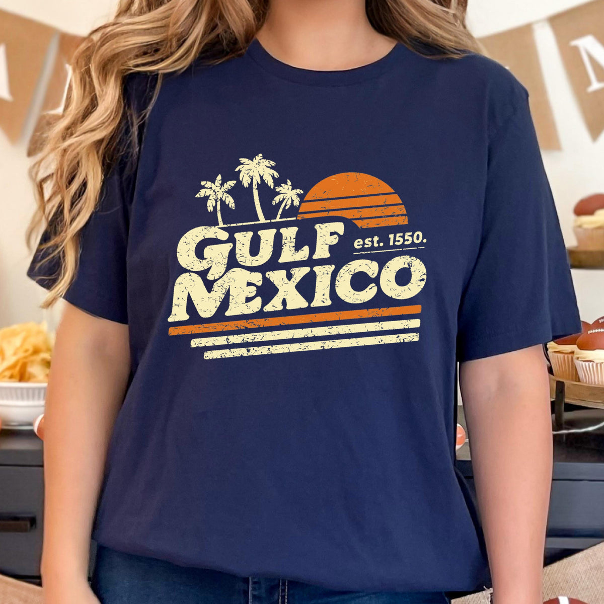 May include: Navy blue t-shirt with a retro design featuring the words "GULF MEXICO" in a distressed font, palm trees, and a setting sun. The text "est. 1550" is also included. The design is in shades of orange and white.