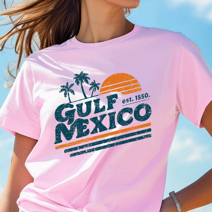 May include: A light pink t-shirt featuring a distressed graphic with the words "GULF MEXICO" in teal, palm trees, and a setting sun. The text "est. 1550" is also included. The shirt has short sleeves.