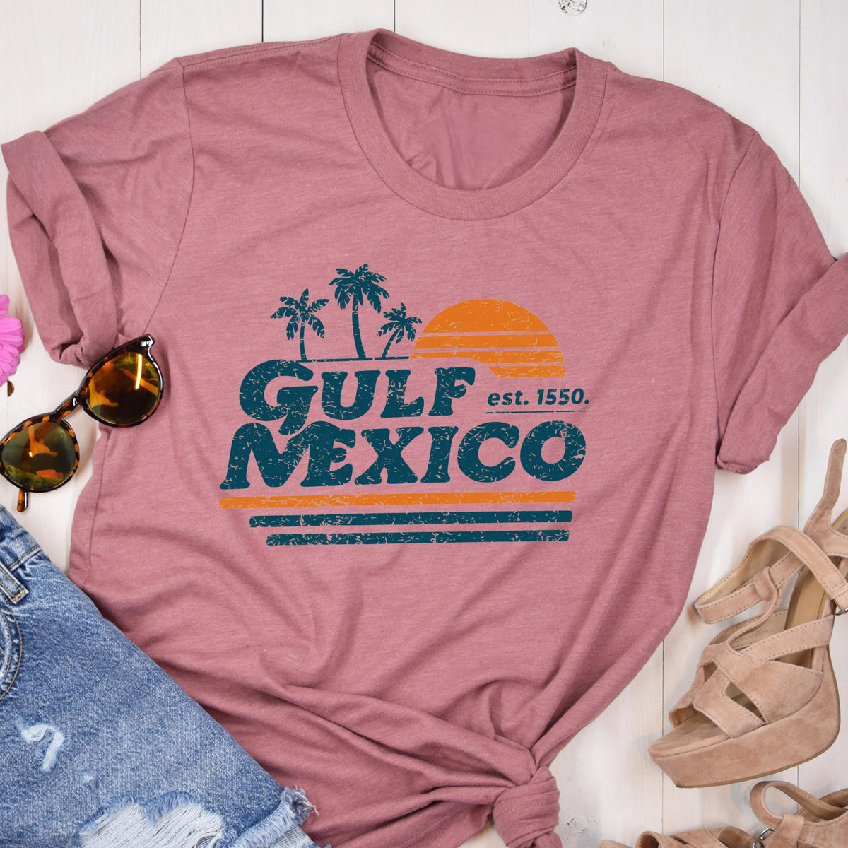 May include: Dusty rose t-shirt with a retro graphic design. The design features the words "GULF MEXICO" in teal, with palm trees, a setting sun, and the text "est. 1550." in orange and teal.