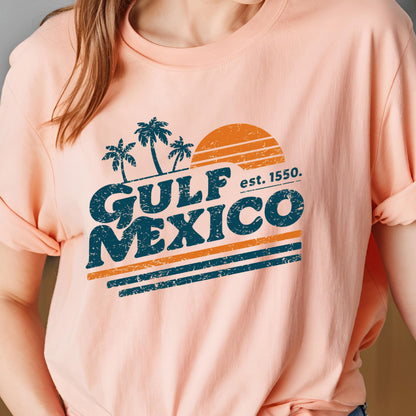 May include: A peach-colored t-shirt featuring a distressed design with the words "GULF MEXICO" in dark teal, with an orange sun and palm trees. The text "est. 1550." is also included. The shirt is a casual wear item.