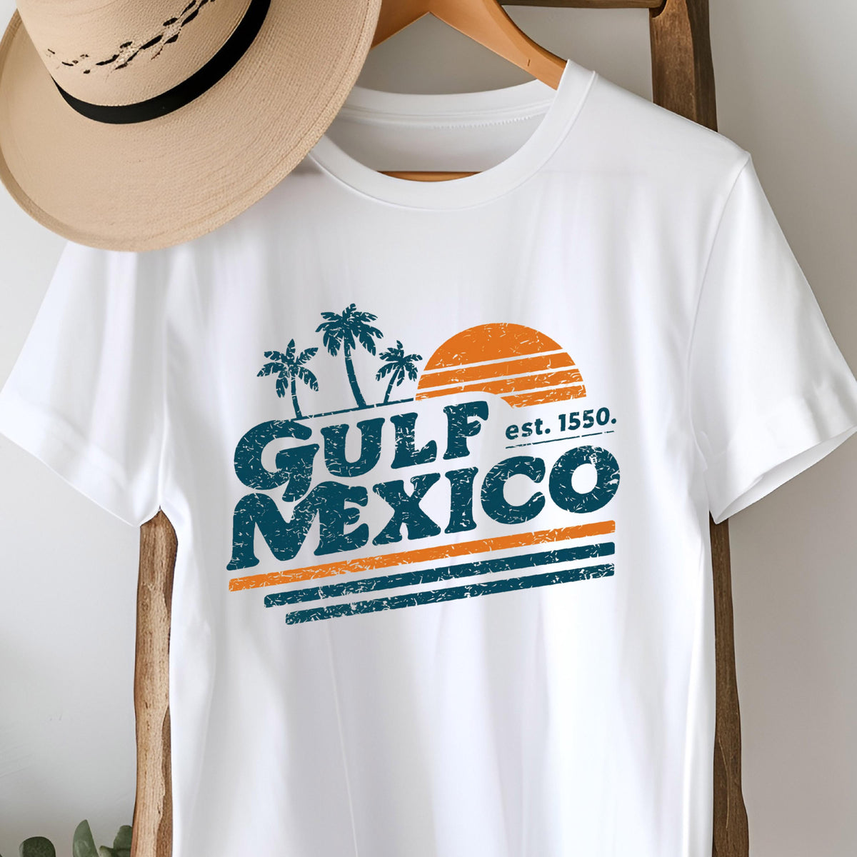 May include: White t-shirt with a retro design featuring the words "GULF MEXICO" in distressed teal lettering, with an orange sun and palm trees. The text "est. 1550" is also included. A tan hat hangs above the shirt.