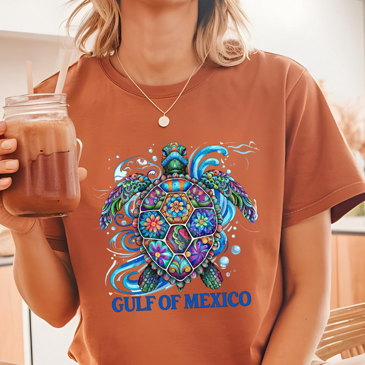 May include: A rust-colored t-shirt featuring a vibrant, multi-colored sea turtle graphic with intricate floral patterns on its shell, surrounded by blue water swirls and bubbles, with the text GULF OF MEXICO printed in bold blue letters below.