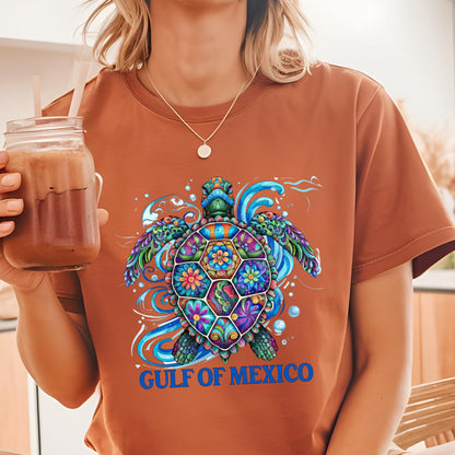 May include: A rust-colored t-shirt featuring a vibrant, multi-colored sea turtle graphic with intricate floral patterns on its shell, surrounded by blue water swirls and bubbles, with the text GULF OF MEXICO printed in bold blue letters below.