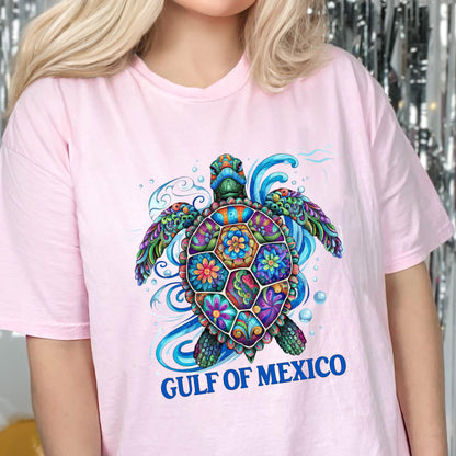 May include: A light pink t-shirt featuring a colorful, intricately patterned sea turtle graphic with floral and geometric designs in shades of blue, purple, green, and orange, surrounded by blue water swirls and the text GULF OF MEXICO.