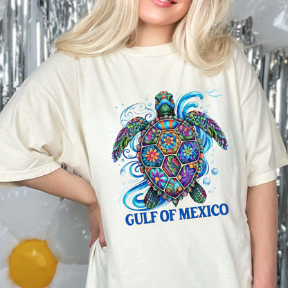 May include: An off-white t-shirt featuring a vibrant, multi-colored sea turtle graphic with intricate floral and geometric patterns on its shell. The design includes swirling blue water elements and the text GULF OF MEXICO in bold blue letters.