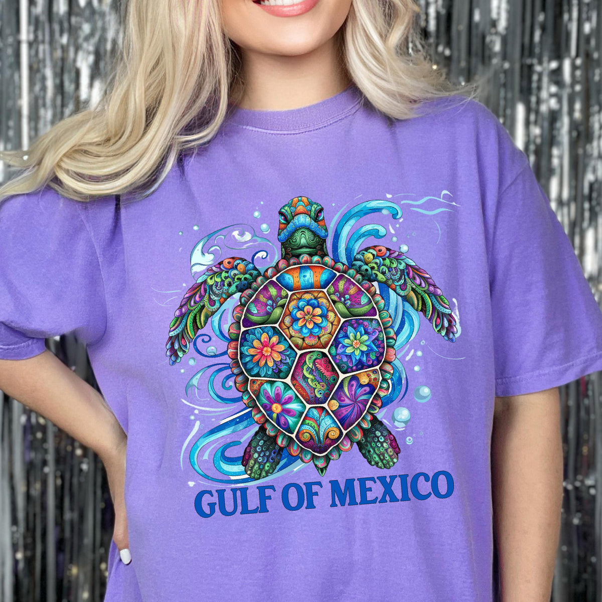 May include: A light purple short-sleeve t-shirt featuring a colorful, intricately patterned sea turtle graphic with floral and swirl designs. Below the turtle, the text GULF OF MEXICO is printed in bold blue capital letters on the front.