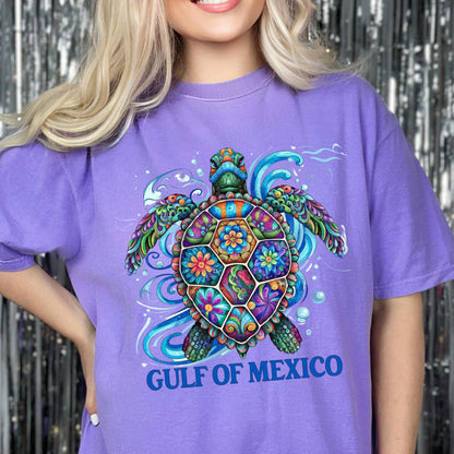 May include: A light purple short-sleeve t-shirt featuring a colorful, intricately patterned sea turtle graphic with floral and swirl designs. Below the turtle, the text GULF OF MEXICO is printed in bold blue capital letters on the front.