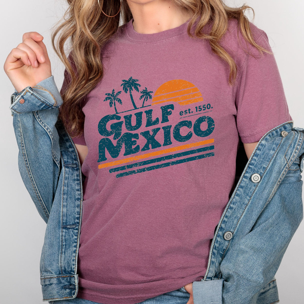 May include: A mauve t-shirt with a graphic design featuring the words "GULF MEXICO" in teal, with an orange sun and palm trees. The text "est. 1550" is also included. The shirt is paired with a denim jacket.