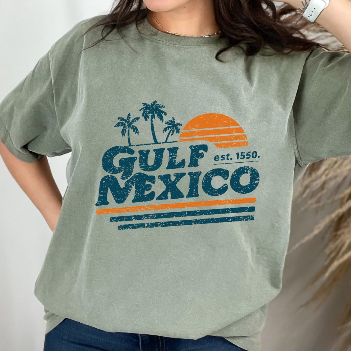 May include: Sage green t-shirt featuring a retro design with the words "GULF MEXICO" in blue, palm trees, and an orange sun. The text "est. 1550" is also included. The design has an aged, vintage look.