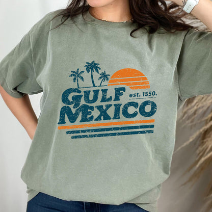 May include: Sage green t-shirt featuring a retro design with the words "GULF MEXICO" in blue, palm trees, and an orange sun. The text "est. 1550" is also included. The design has an aged, vintage look.