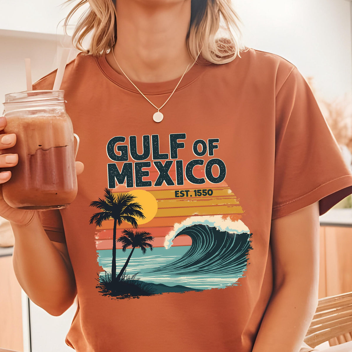 May include: A brown t-shirt with a retro-style graphic of a sunset, palm trees, and a wave. The text "GULF OF MEXICO EST. 1550" is printed on the shirt.