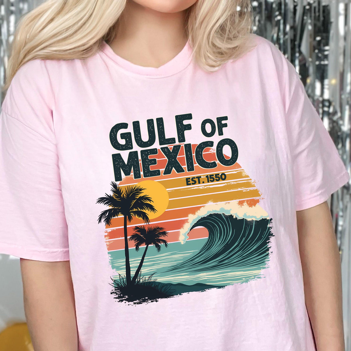 May include: Pink t-shirt with a graphic design of a wave, palm trees, and a sunset. The text "GULF OF MEXICO EST. 1550" is printed on the shirt.