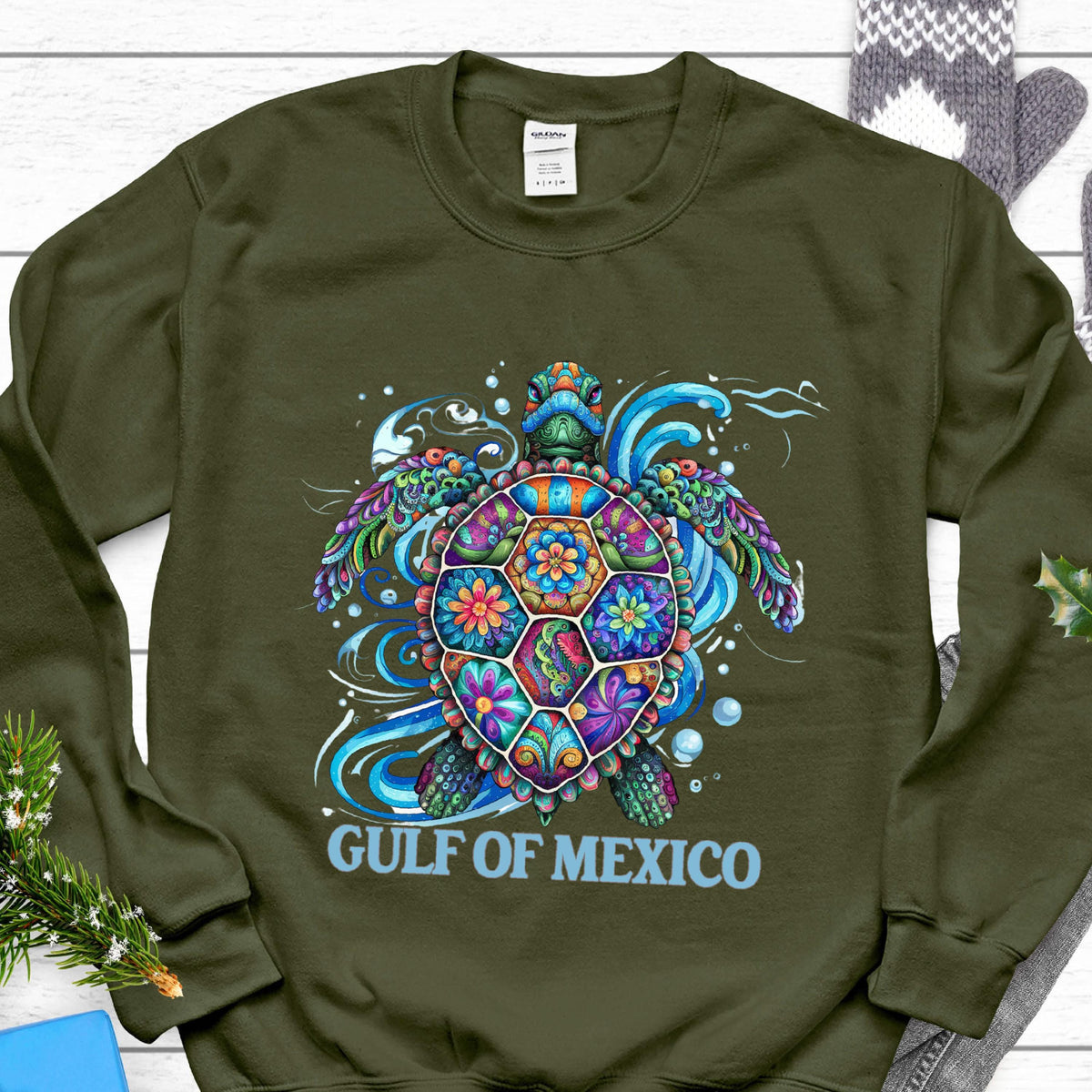 May include: An olive green sweatshirt featuring a colorful sea turtle design. The turtle's shell is decorated with floral patterns in shades of blue, orange, purple, and green. The words "GULF OF MEXICO" are printed in light blue below the turtle.