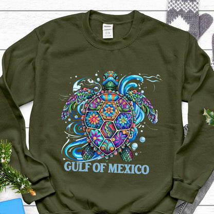 May include: An olive green sweatshirt featuring a colorful sea turtle design. The turtle's shell is decorated with floral patterns in shades of blue, orange, purple, and green. The words "GULF OF MEXICO" are printed in light blue below the turtle.