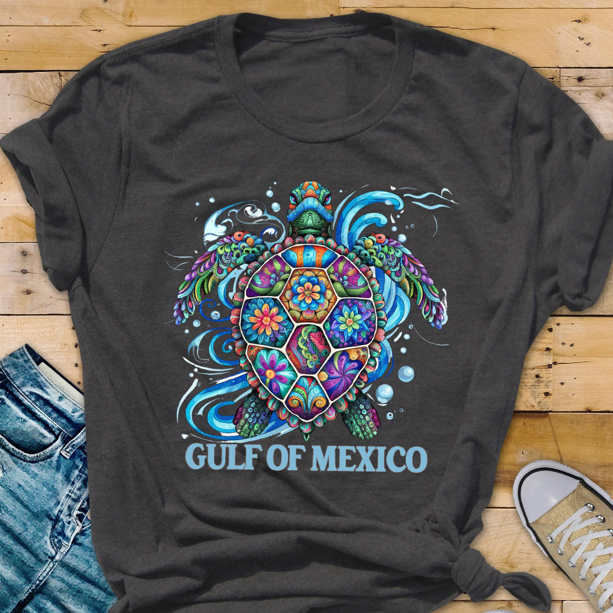 May include: A dark gray t-shirt with a colorful, floral patterned sea turtle design. The turtle is surrounded by blue waves and bubbles. The text "GULF OF MEXICO" is printed in light blue below the turtle.