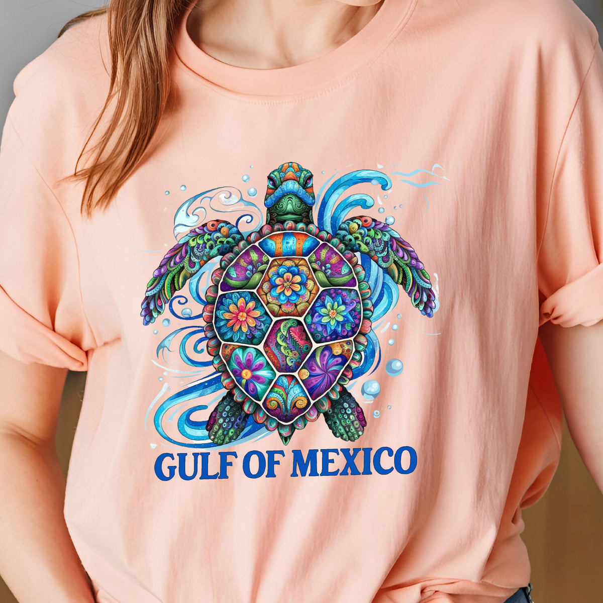 May include: A light pink t-shirt with a colorful, patterned sea turtle design. The turtle is surrounded by blue waves and bubbles. The text "GULF OF MEXICO" is printed in blue below the turtle.