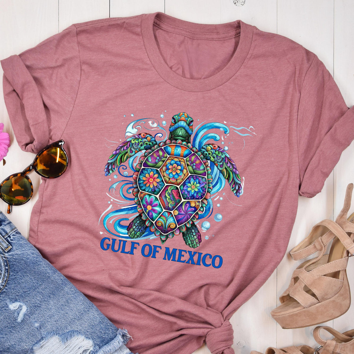 May include: A pink t-shirt with a colorful, floral patterned sea turtle design. The text "GULF OF MEXICO" is printed in blue below the turtle.