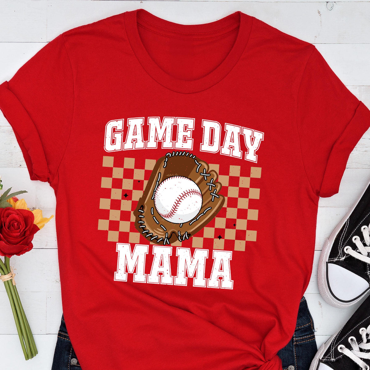 May include: A bright red short-sleeved t-shirt featuring a graphic print with the text GAME DAY MAMA in white block letters. The design includes a brown baseball glove holding a white baseball, centered over a tan and brown checkered background pattern.