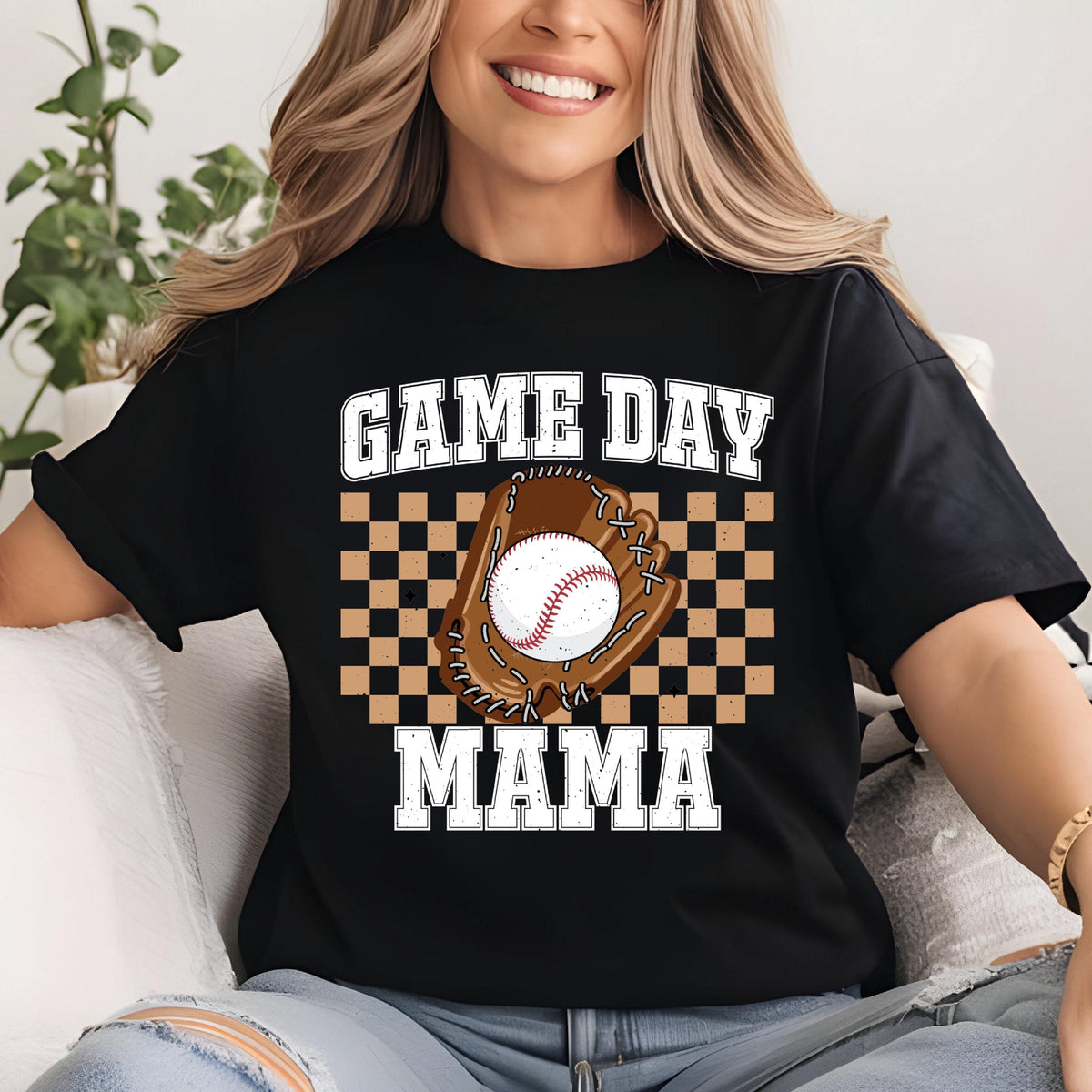 May include: A black short-sleeved t-shirt featuring a graphic print with the text GAME DAY MAMA in white block letters. The design includes a brown baseball glove holding a white baseball, centered over a tan and black checkered square pattern.
