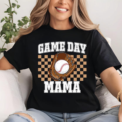 May include: A black short-sleeved t-shirt featuring a graphic print with the text GAME DAY MAMA in white block letters. The design includes a brown baseball glove holding a white baseball, centered over a tan and black checkered square pattern.